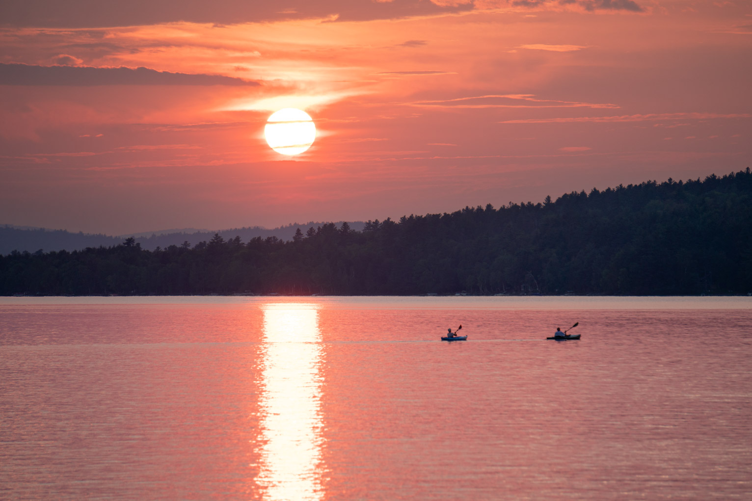 The sun starts to sink behind the horizon clouds over the sunset lit lake where there are two kayakers